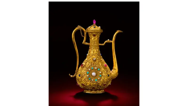 A gold ewer encrusted with gemstones and pearls, photographed against a dark red background.