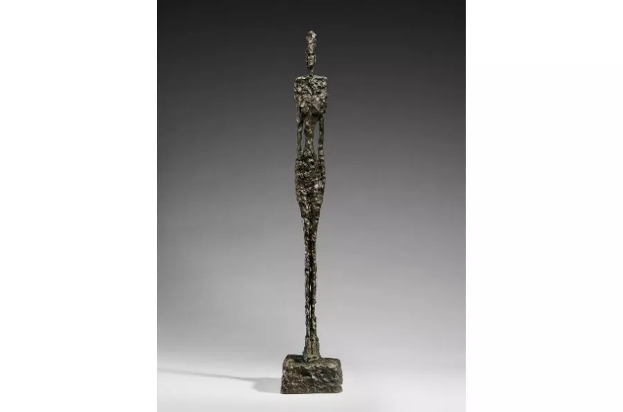 A tall, thin bronze sculpture of a standing human figure on a small base, set against a gray background.