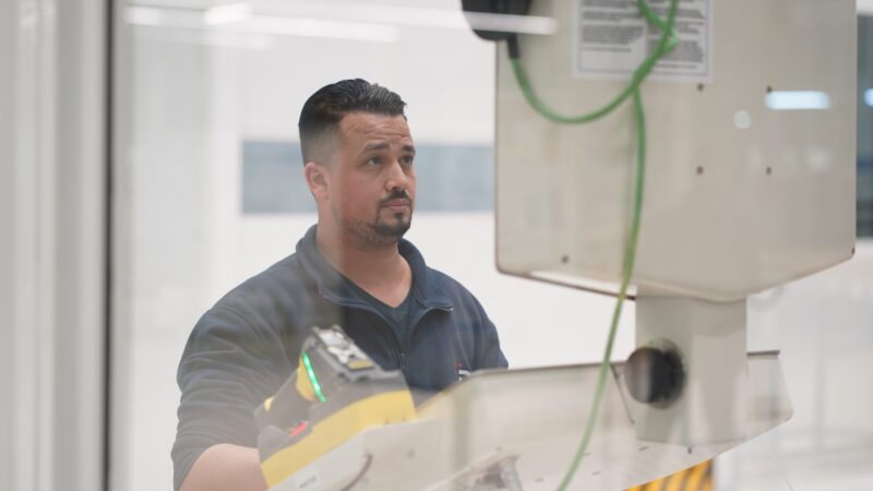 A man in a workshop uses a handheld tool while looking up at large industrial equipment.