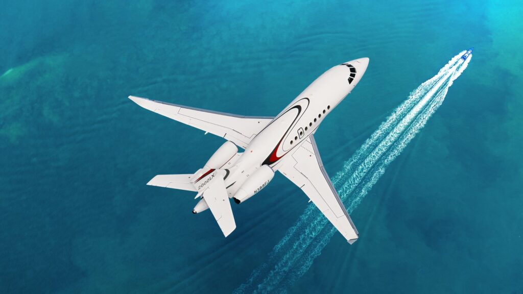 A white Dassault Falcon 2000LX business jet flies over turquoise ocean water near a speedboat leaving a wake.