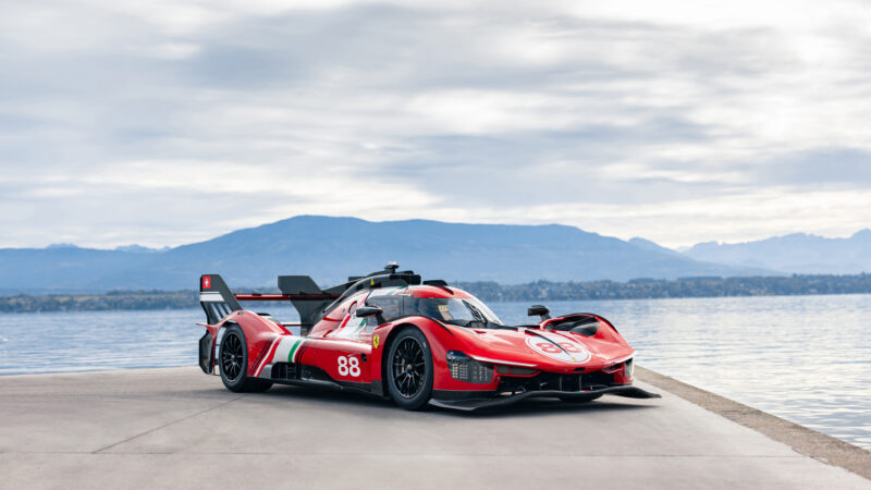 A red Ferrari 499P Modificata race car with number 88 parked on a dock beside a lake and mountains.
