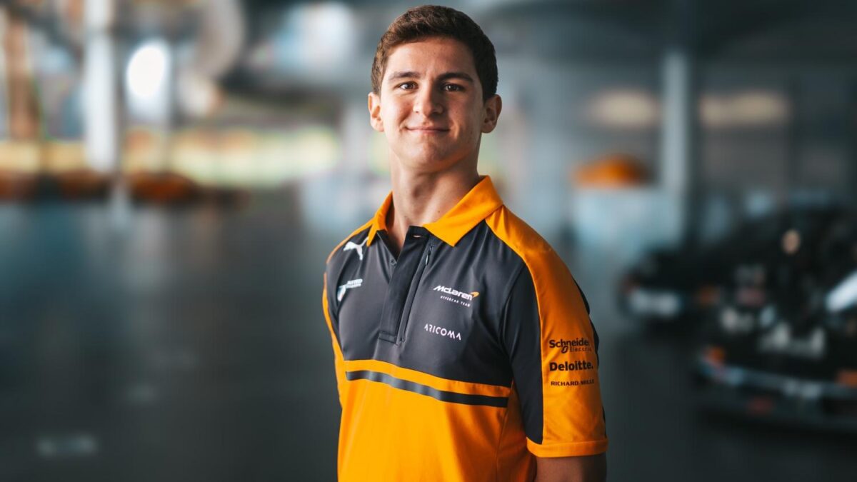 A young man in an orange and black McLaren team polo shirt stands smiling in a garage with race cars blurred behind him.