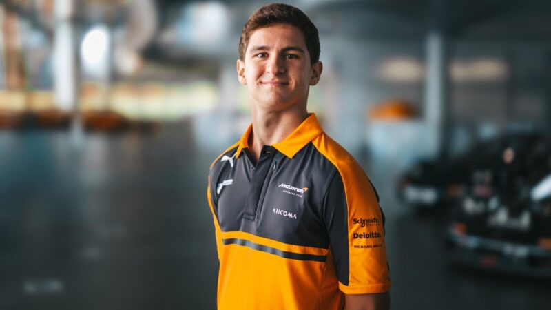 A young man in an orange and black McLaren team polo shirt stands smiling in a garage with race cars blurred behind him.
