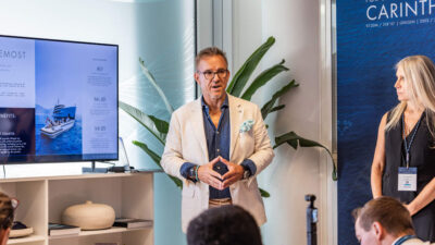 A man in a blazer speaks at a press event in front of screens showing Fraser Yachts charter information and a yacht banner.