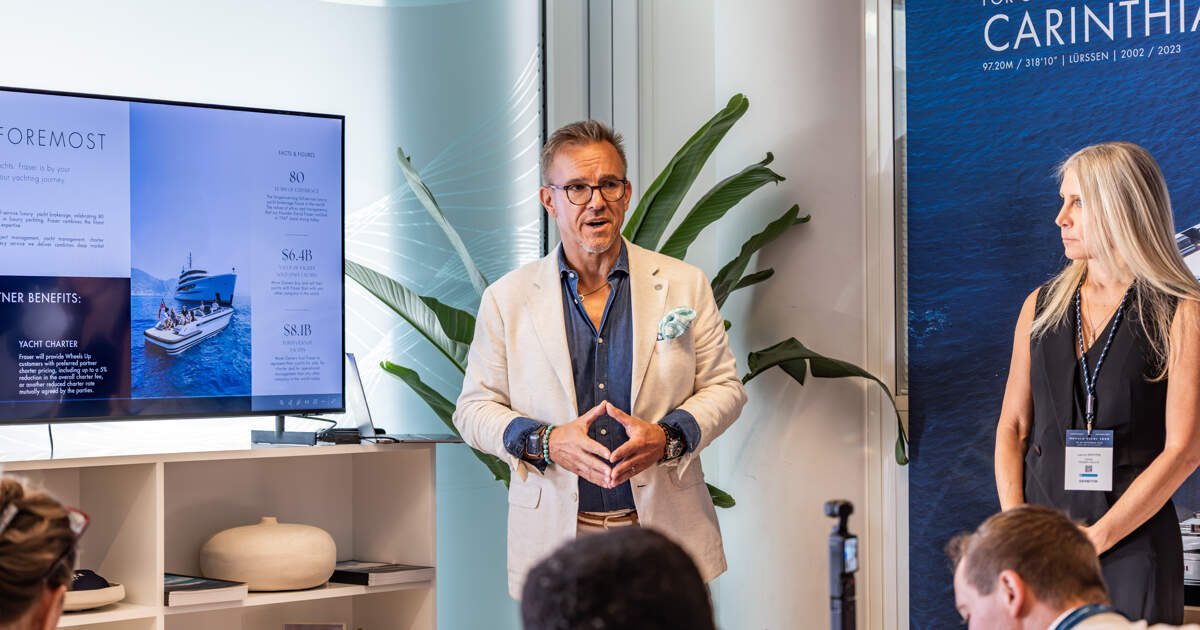A man in a blazer speaks at a press event in front of screens showing Fraser Yachts charter information and a yacht banner.