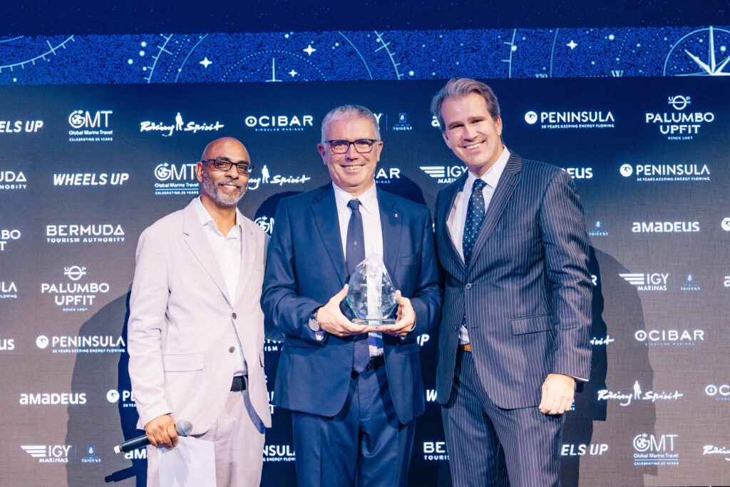 Three men in suits pose on stage with a glass trophy in front of a backdrop covered in sponsor logos.