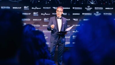 A man in a tuxedo speaks on stage holding papers in front of a backdrop of sponsor logos including Bermuda Tourism Authority and Wheels Up.