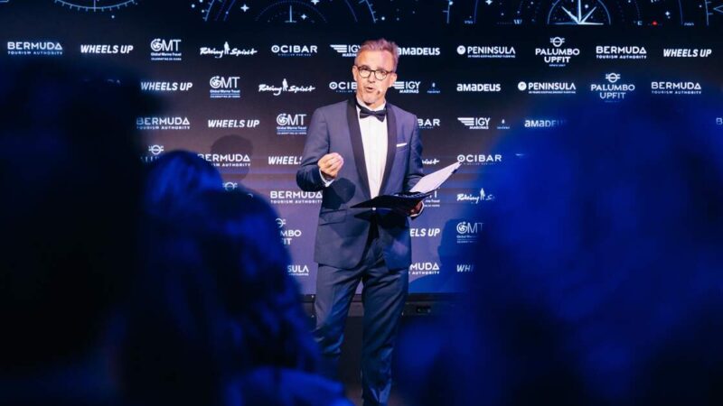 A man in a tuxedo speaks on stage holding papers in front of a backdrop of sponsor logos including Bermuda Tourism Authority and Wheels Up.