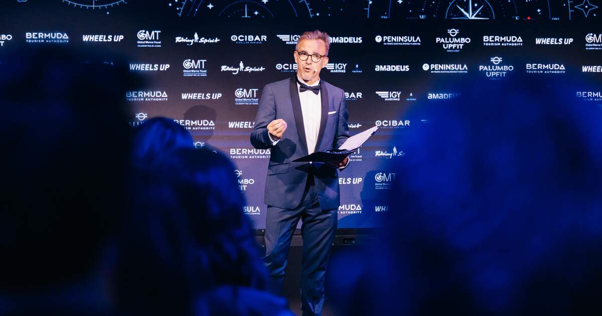 A man in a tuxedo speaks on stage holding papers in front of a backdrop of sponsor logos including Bermuda Tourism Authority and Wheels Up.