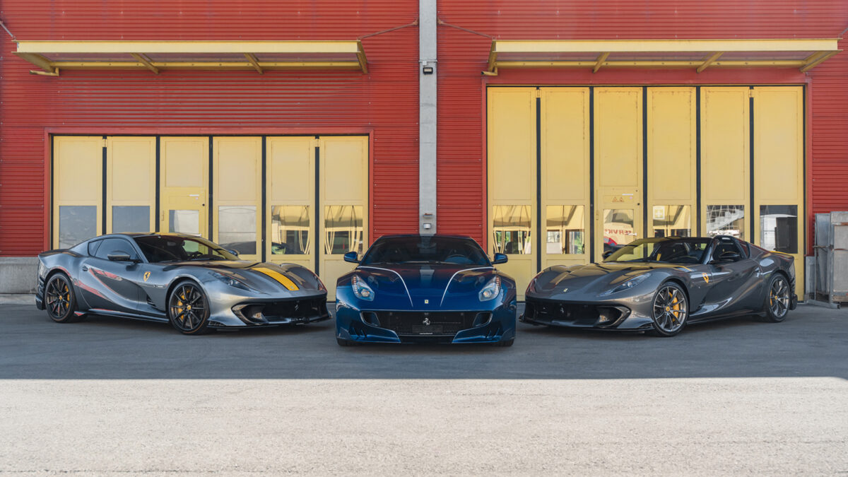 Three Ferrari sports cars, silver, blue, and gray, parked side by side in front of a red building with yellow doors.