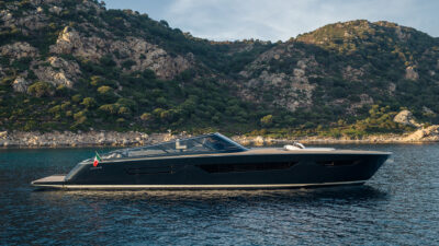 A dark-hulled open yacht with an Italian flag anchored near a rocky, green coastline.