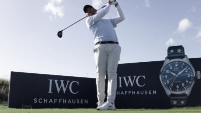 A golfer swings a club on a course in front of IWC Schaffhausen branded boards and a large watch graphic.