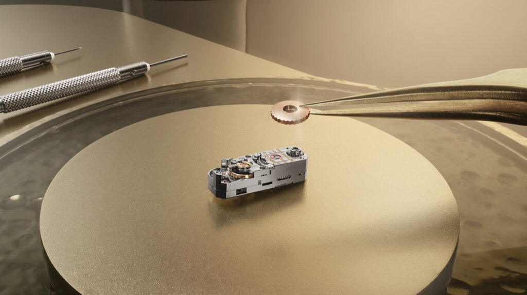 Close-up of a miniature watch movement on a gold surface with tweezers holding a small gear component above it.