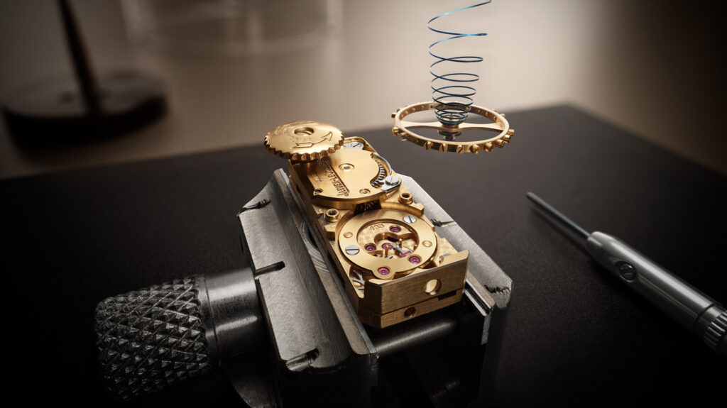Close-up of a gold watch movement with a balance wheel and blue hairspring suspended above it, held in a watchmaker's vice.