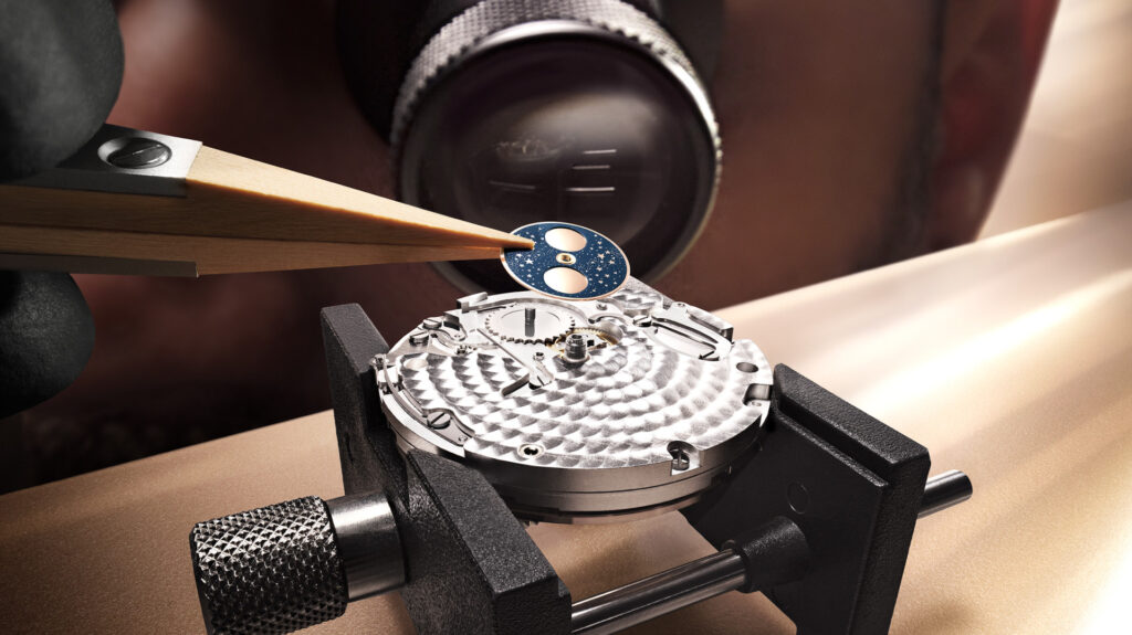 Tweezers placing a blue moon-phase disc onto a watch movement held in a workbench vice.
