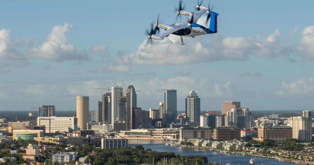 A Joby eVTOL aircraft flies above a Florida city skyline with a river below.