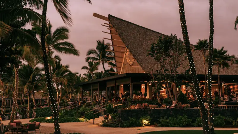 Twilight view of a thatched-roof resort pavilion surrounded by string-lit palm trees and a stone wall.