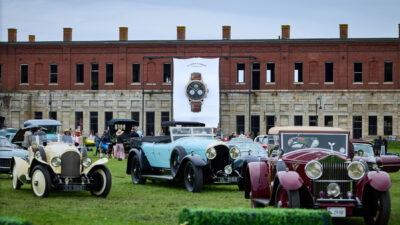 Vintage classic cars parked on a lawn before a brick fort building with an A. Lange & Söhne watch banner hanging between them.