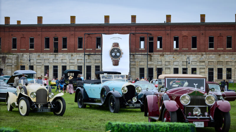 Vintage classic cars parked on a lawn before a brick fort building with an A. Lange & Söhne watch banner hanging between them.