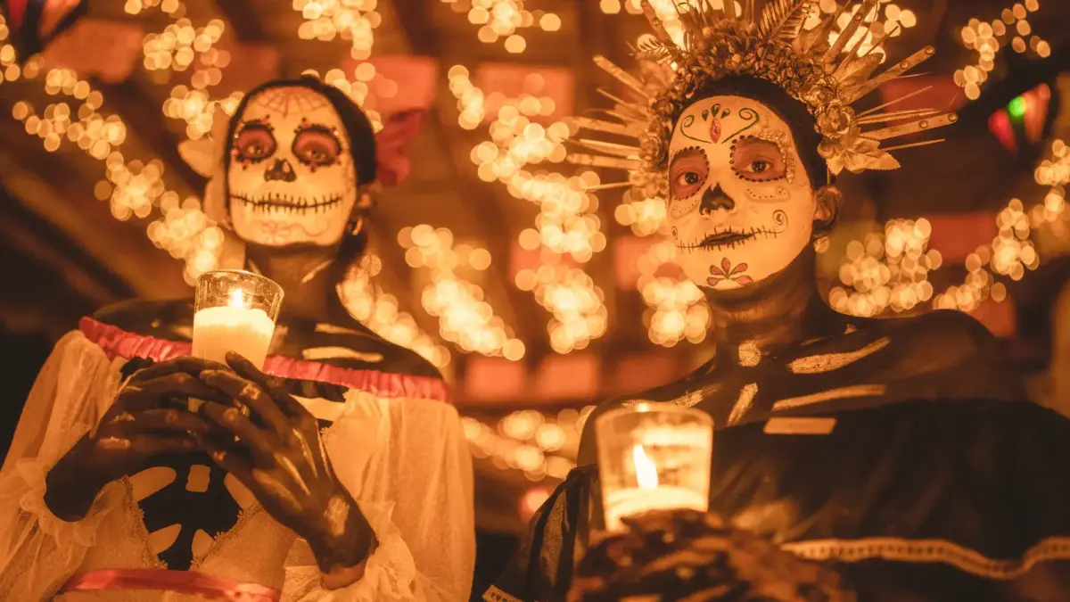 Two people in Día de Muertos skull makeup and costumes hold lit candles amid warm, twinkling lights.