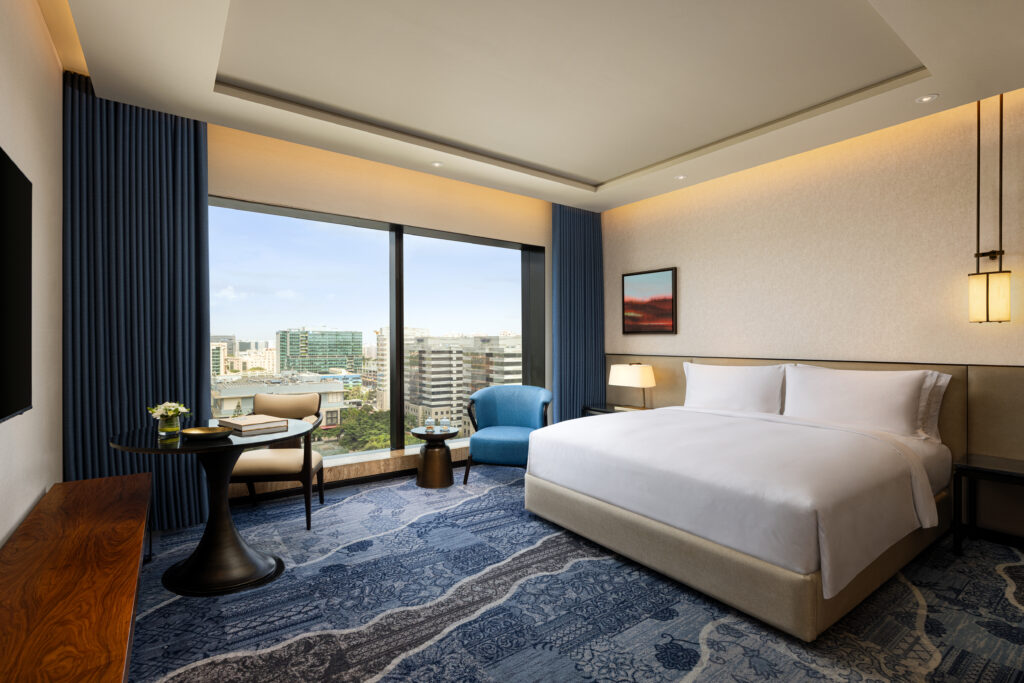 Modern hotel room with a large bed, blue armchair, round table, and city skyline view through a window.