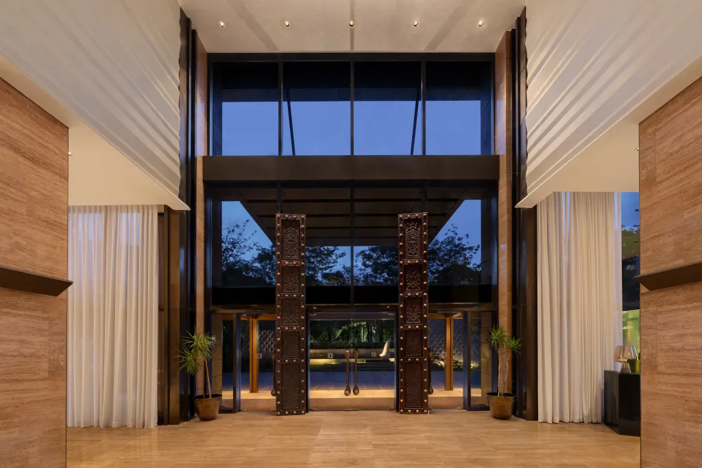Hotel lobby entrance with tall glass doors, carved wooden pillars, marble floors, and potted plants on each side.