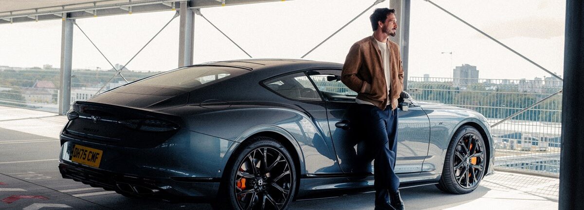 A man in a tan jacket stands beside a dark blue Bentley coupe in a multi-level parking structure overlooking a city.