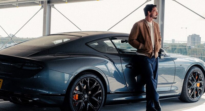 A man in a tan jacket stands beside a dark blue Bentley coupe in a multi-level parking structure overlooking a city.