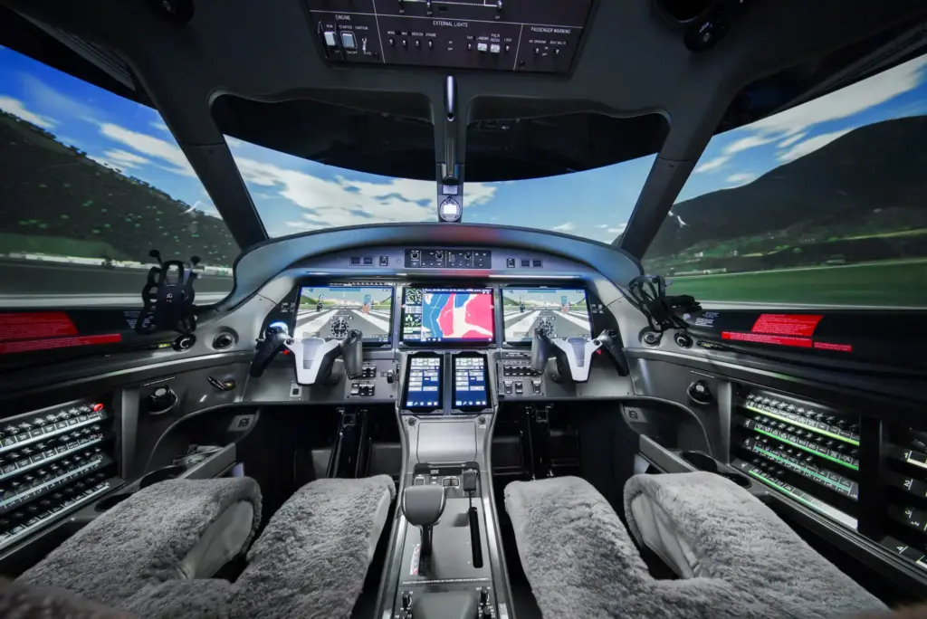 Interior view of an aircraft simulator cockpit with dual control yokes, multiple digital avionics screens, and a projected outdoor landscape visible through the