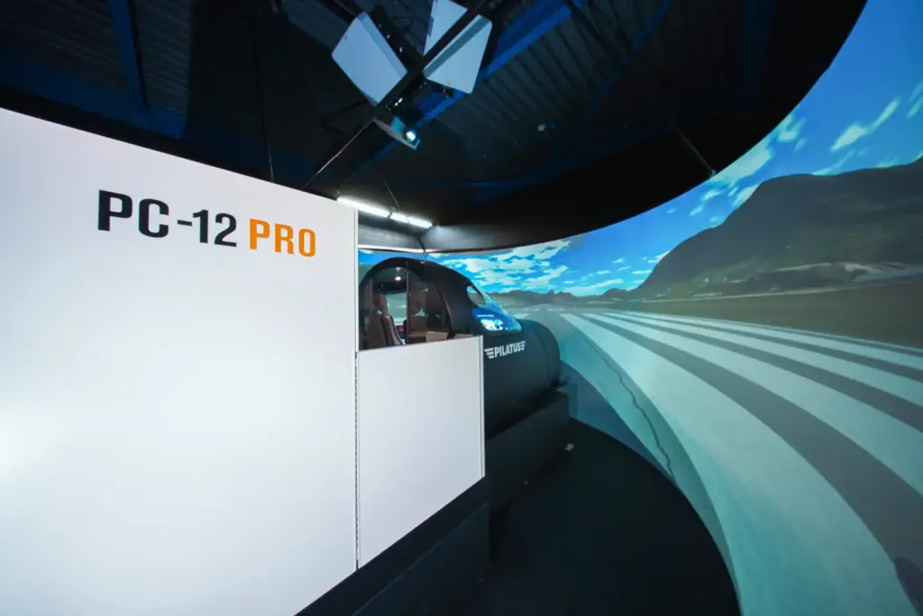 A Pilatus PC-12 PRO flight simulator pod with a curved screen showing a mountainous runway scene.