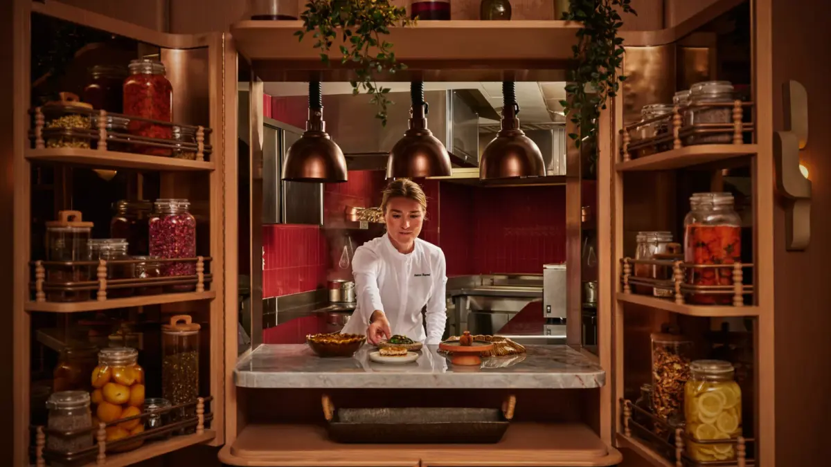 A chef in whites plates food at a marble counter framed by wooden cabinets filled with jars of preserves and spices.