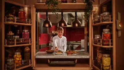 A chef in whites plates food at a marble counter framed by wooden cabinets filled with jars of preserves and spices.