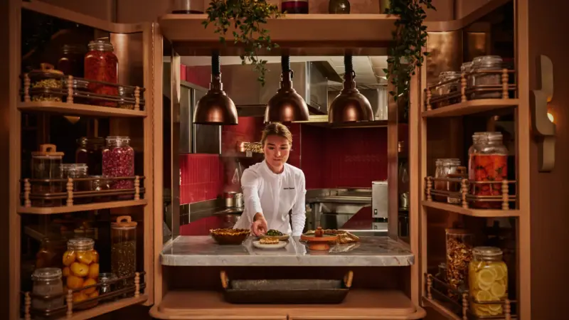 A chef in whites plates food at a marble counter framed by wooden cabinets filled with jars of preserves and spices.