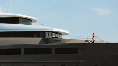 Two people stand on the upper deck of a modern superyacht against a clear sky.