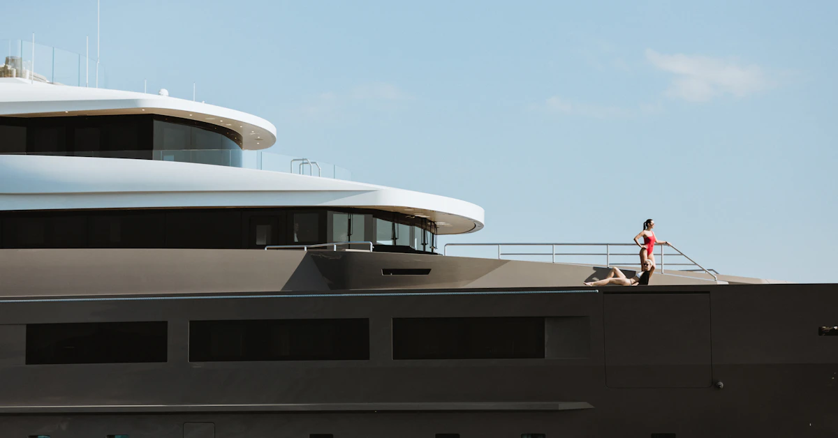 Two people stand on the upper deck of a modern superyacht against a clear sky.