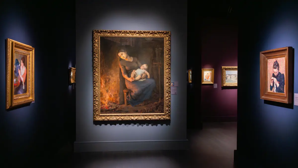 A gallery hallway with illuminated framed paintings, including a central portrait of a woman holding an infant by a fireplace.