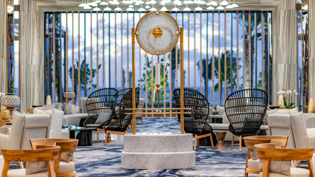A hotel lobby lounge features a large gold and marble pendulum clock surrounded by wicker chairs with an ocean view.