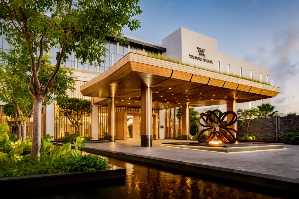 Entrance of the Waldorf Astoria Riviera Maya with a wooden canopy, illuminated sculpture, and greenery at dusk.