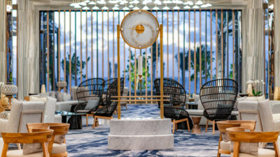 A hotel lobby lounge features a large gold and marble pendulum clock surrounded by wicker chairs with an ocean view.